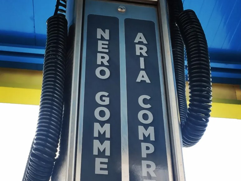 Colonnina in acciaio con scritte NERO GOMME e ARIA COMPR, tubi neri a spirale sotto tettoia blu e gialla